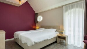 A modern hotel room with a large bed, a bedside table with a lamp, a round wall clock, and a window with sheer curtains. - Holiday Home in Oostkapelle Near Beach (Oostkapelle)