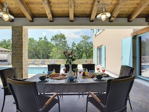Zona pranzo all'aperto con vista piscina, colonna in pietra e travi in legno al soffitto.