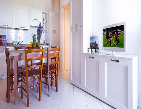 A modern kitchen with white cabinets, a dining table with wooden chairs, and a flat-screen TV mounted on a white cabinet. - Charming Chianti Retreat (Barberino Tavarnelle)