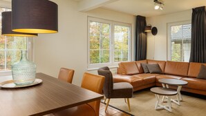 Una sala de estar moderna con un sofá de cuero marrón, una mesa de comedor y sillas. Hay dos ventanas con cortinas, y una lámpara cuelga sobre la mesa.