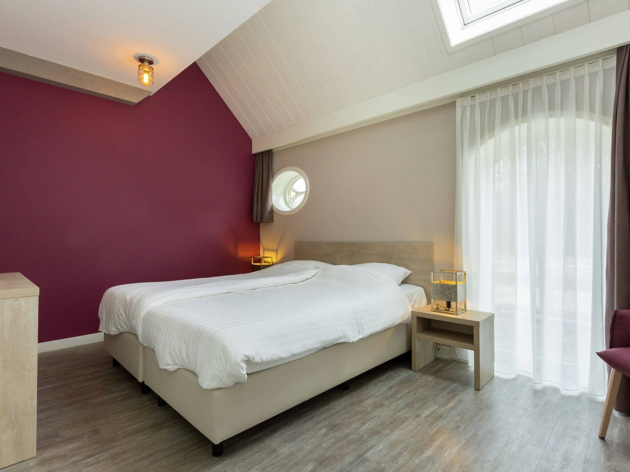 Une chambre d’hôtel moderne avec un grand lit, une table de chevet avec une lampe, une horloge ronde fixée au mur et une fenêtre avec des rideaux légers.