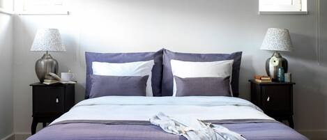 Una cama bien hecha con una colcha morada, dos mesitas de noche con lámparas, y una ventana con vistas.