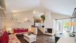 Amplia sala de estar con sofá, una mesa de centro y chimenea. Se observa una cocina al fondo con una escalera que sube.