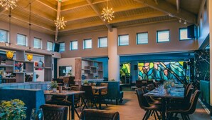 A spacious restaurant with high ceilings, wooden beams, and a central bar area. Tables are set with chairs, and there are potted plants on the tables. - Comfortable Chalet in the Green Kempen (Mol)