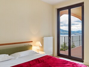 A hotel room with a bed, a nightstand with a lamp, and a large sliding glass door leading to a balcony with a view of the mountains and a lake. - Residential Complex With Panoramic Views and Pool (Oggebbio)