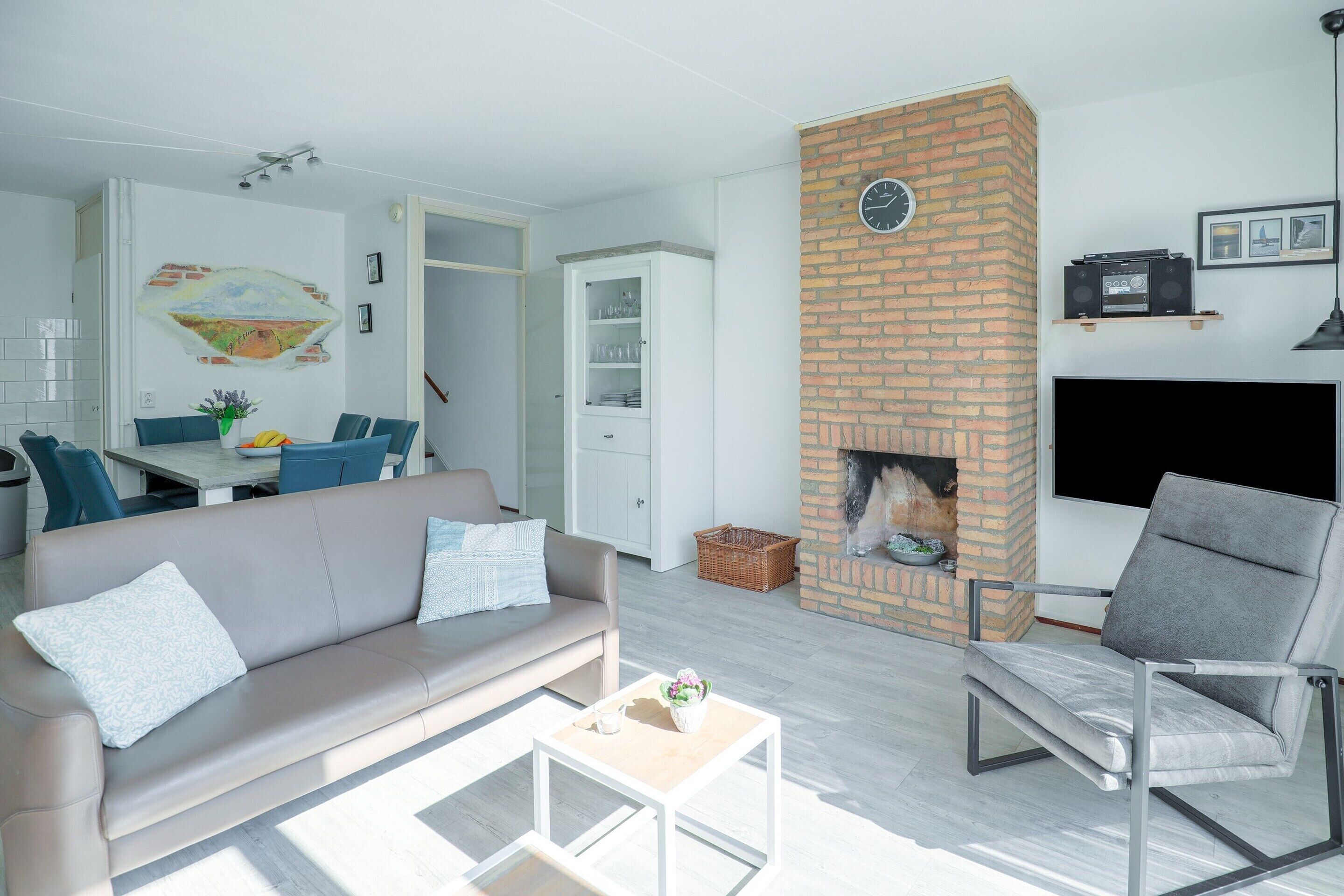 A modern living room with a sofa, armchair, and coffee table. A fireplace is built into a brick wall, and there is a television mounted above it.