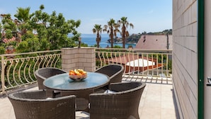 A balcony with wicker furniture, a table with a fruit bowl, and a view of the sea and palm trees. - Villa in Sumartin Near the Beach & Ferry Port (Selca)