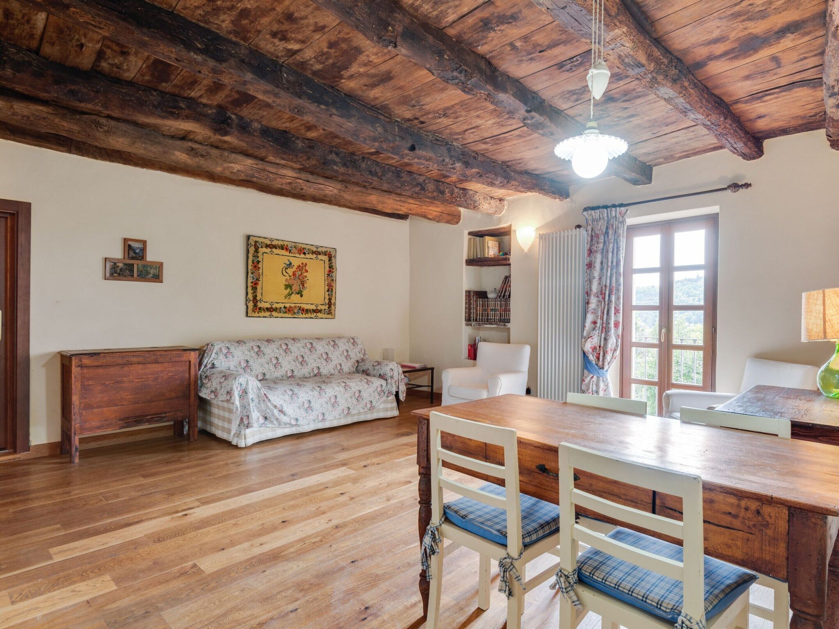 A rustic room with a wooden ceiling, a floral sofa, a wooden dining table with chairs, a bookshelf, and a window with curtains.