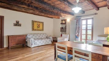 Ein rustikaler Raum mit Holzbalkendecke, einem blumengemusterten Sofa, einem Holzesstisch mit Stühlen, einem Bücherregal und einem Fenster mit Vorhängen.