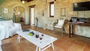 Un vaste espace de vie agrémenté d’un mur en pierre, d’un canapé blanc, d’un fauteuil en osier, d’une table basse avec des verres à vin et d’un téléviseur à écran plat fixé au mur.