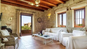 Une pièce chaleureuse avec des murs en pierre, des poutres apparentes, un canapé blanc, un fauteuil en osier et une table avec des boissons.