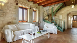Un vaste espace de vie doté d’un plafond en bois, de murs en pierre, d’un canapé blanc, d’une table à manger avec des verres à vin et un panier de fruits, ainsi que d’un escalier vert.