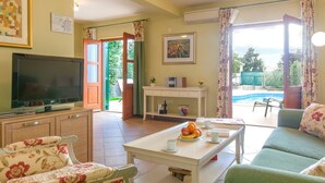 A living room with a television, floral chairs, a white table with fruit, and a view of a swimming pool. - Charming Villa With Spacious Rooms and Private Swimming Pool in the Lavender Island of Hvar (Hvar)