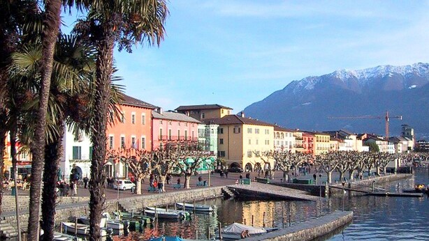 A waterfront promenade with buildings, palm trees, and a mountain in the background. - Residential Complex With Panoramic Views and Pool (Oggebbio)