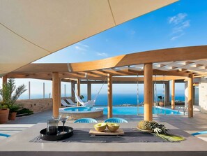 An outdoor pool area with a view of the sea, wooden pergolas, lounge chairs, and a table with fruit and a drink tray.