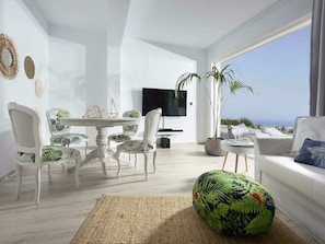 A modern living room with a white color scheme, a round table, and chairs with green leaf patterns. There is a large flat-screen TV mounted on the wall, a potted plant, and a view of the ocean through a large window. - Luxury Villa With Heated Pool in Agia Pelagia (Malevizi)