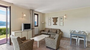A living room with a sofa, two armchairs, a coffee table, a TV, and a dining table with chairs. There are framed pictures on the wall and a view of the outdoors through a window. - Residential Complex With Panoramic Views and Pool (Oggebbio)