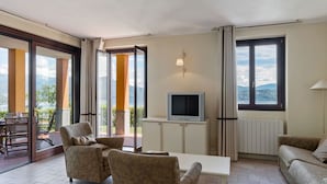 A spacious living room with a sofa, armchairs, a TV, and a table. Large windows offer a view of the outdoors. - Residential Complex With Panoramic Views and Pool (Oggebbio)
