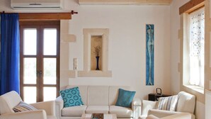 A living room with a white sofa, a coffee table, and a TV. There is a window with curtains, a wall-mounted air conditioner, and a shelf with decorative items. - Dreamy Villa in Rethymnon With Private Pool (Rethymno)
