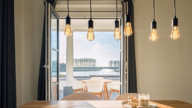 A dining area with a wooden table, hanging light fixtures, and a view of a body of water and trees through open doors. - Modern Apartment 2 Bath Rooms, Close to Lake Veere (Kamperland)
