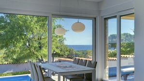 A modern dining area with a table and chairs, large windows with a view of the sea and greenery, and a hanging light fixture. - Villa in Peroj With Pool Near Barbariga Beach (Vodnjan)