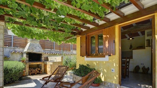 Une terrasse extérieure dotée d’une cheminée en pierre, de fauteuils de jardin en bois et d’une pergola recouverte de vignes.