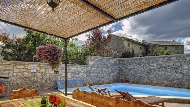 Un espace piscine extérieur avec des murs en pierre, une table en bois entourée de chaises et une pergola au-dessus.