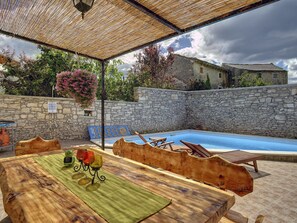 Et udendørs poolområde med stenvægge, et træbord med stole og en pergola over hovedet.