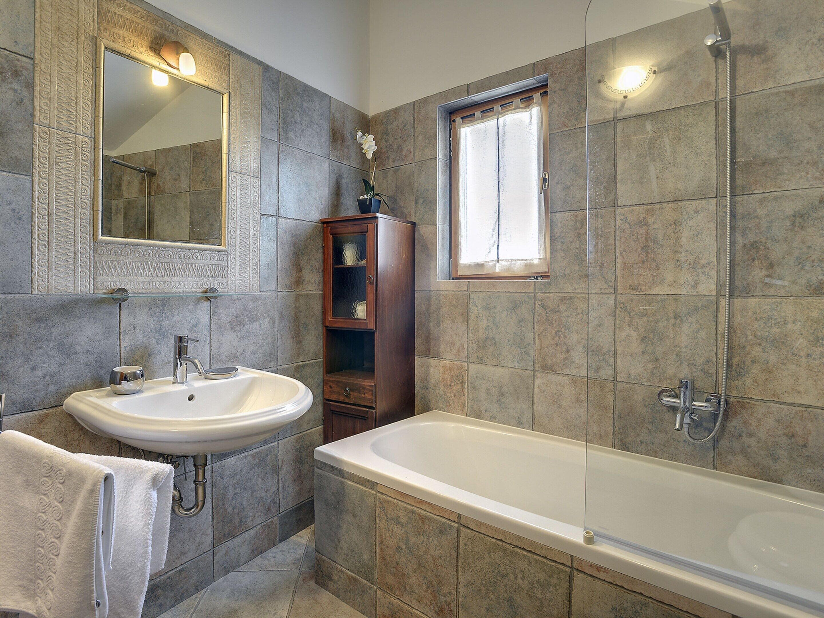 Un baño con bañera, lavamanos y ducha. Hay una ventana sobre la ducha y un espejo sobre el lavamanos.