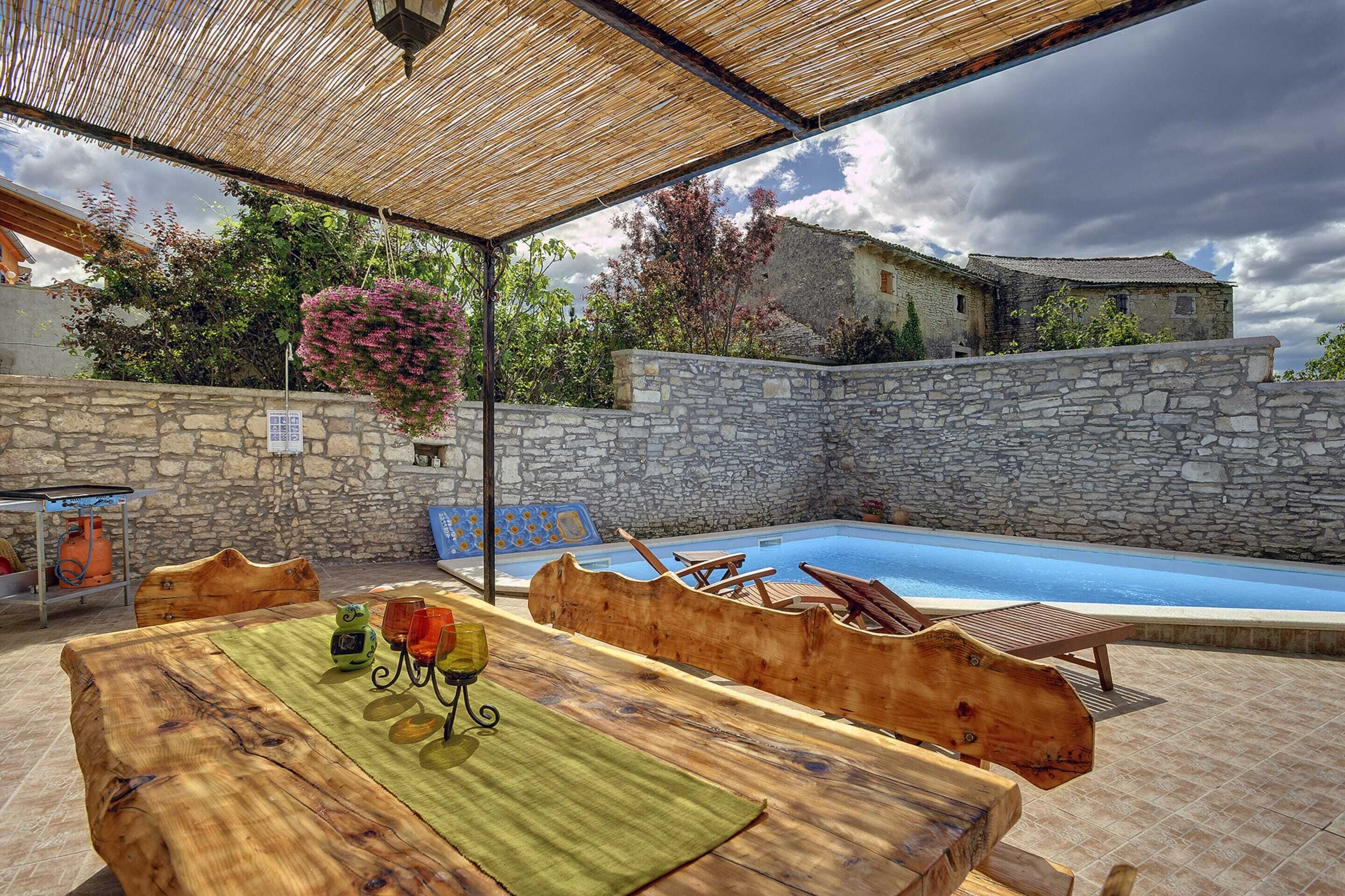 An outdoor pool area with stone walls, wooden furniture, and a covered seating area.
