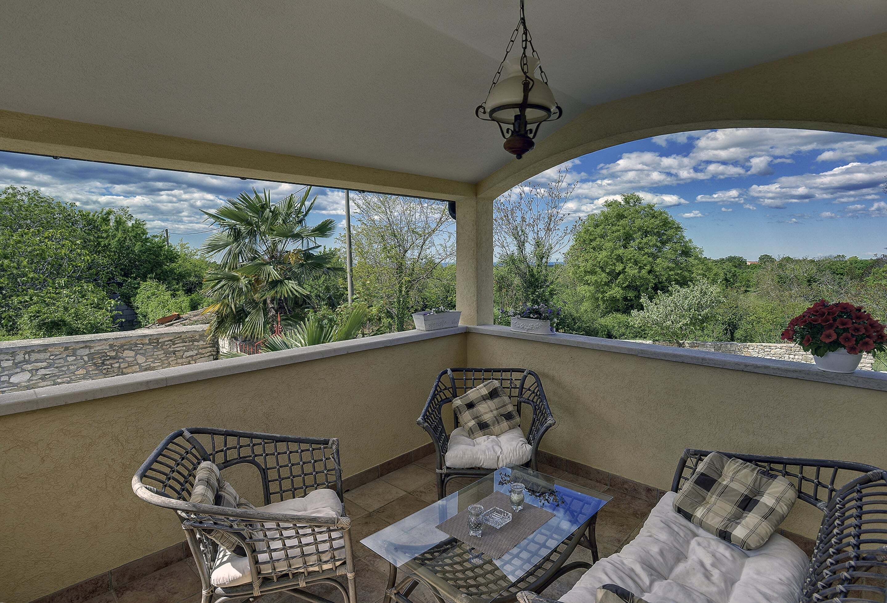 A covered outdoor seating area with wicker chairs and a glass table, overlooking a lush landscape with trees and a clear sky.