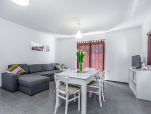 Un moderno salón con un sofá gris, una mesa de comedor blanca y sillas. Hay una pequeña zona de cocina con un televisor y una máquina de café.