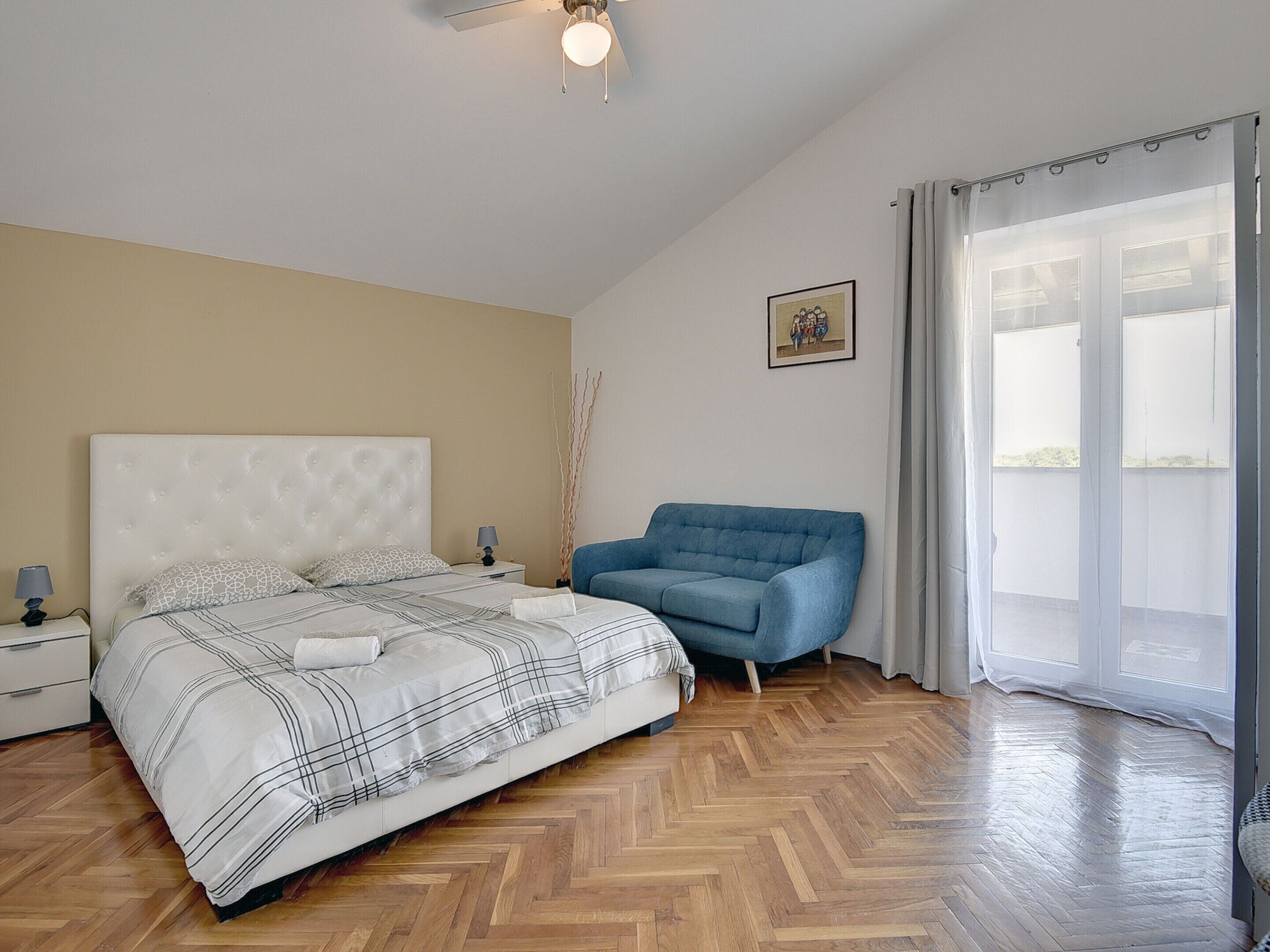 Ein Schlafzimmer mit einem Bett, einem blauen Sofa, einem Nachttisch, einer Lampe, einem Bild an der Wand und einem Fenster mit Vorhängen.