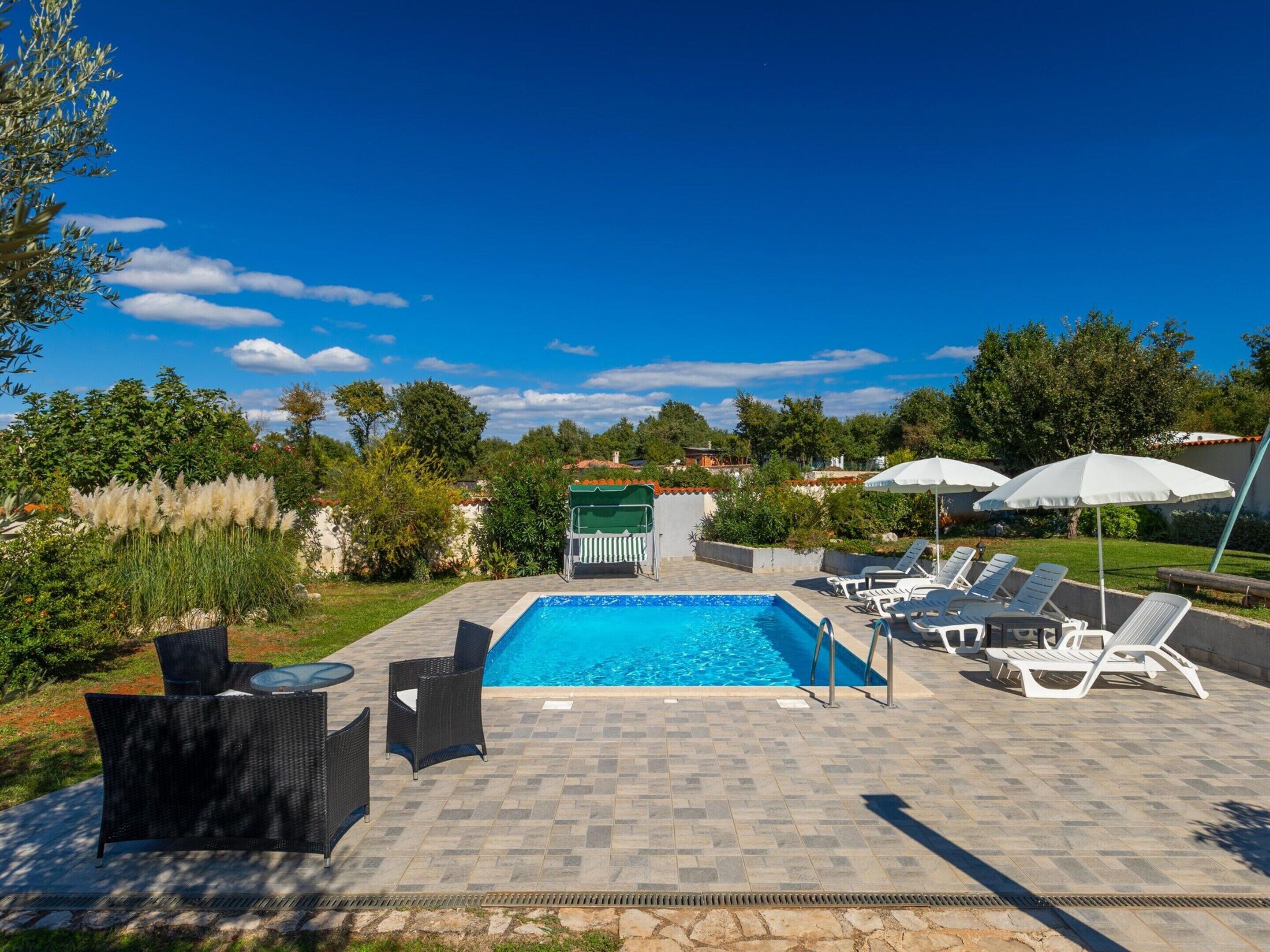 Une piscine entourée de verdure, avec des chaises longues et des parasols, sous un ciel dégagé.