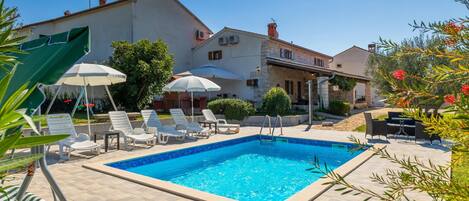 A swimming pool with lounge chairs and umbrellas, surrounded by stone buildings and greenery.
