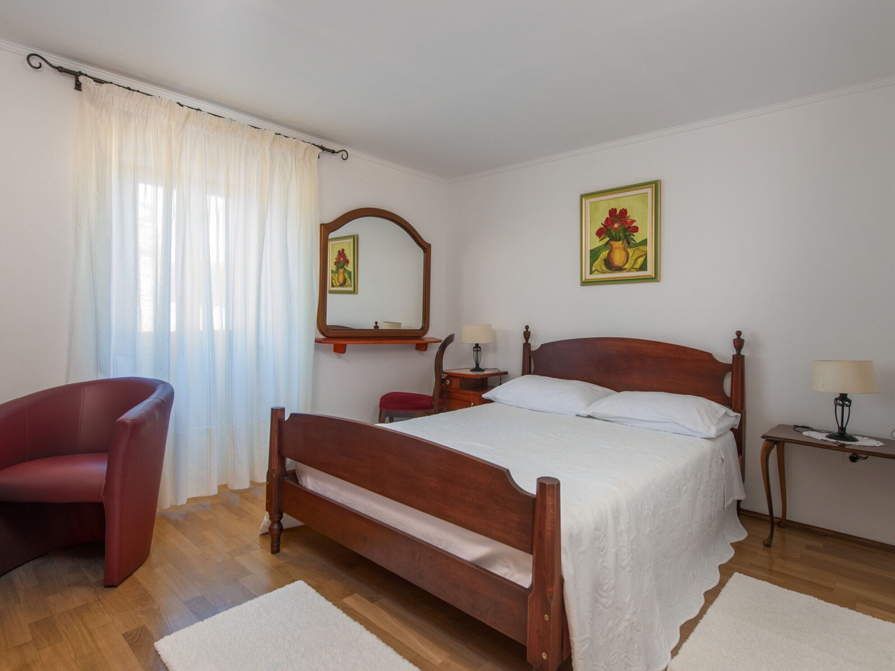 A bedroom with a wooden bed, a red chair, a bedside table with a lamp, a mirror, and a painting on the wall.