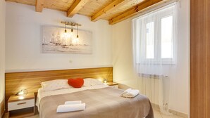 Una camera da letto con soffitto in legno, un letto con copriletto color beige, un cuscino a forma di cuore rosso, due asciugamani bianchi, un comodino con una lampada e un quadro raffigurante barche a vela.