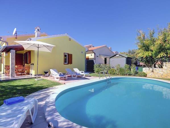 A swimming pool with lounge chairs and a table, surrounded by a stone wall and greenery.