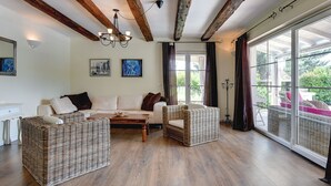 Amplia sala de estar con vigas de madera a la vista, un ventanal con vistas a la vegetación, un conjunto de sofás y mobiliario de mimbre.