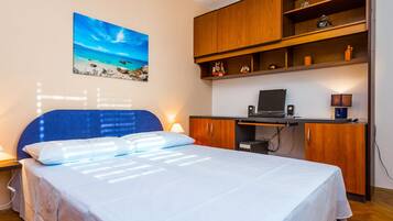 A bedroom with a bed, a desk with a laptop, a shelf with decorative items, and a painting of a beach scene.