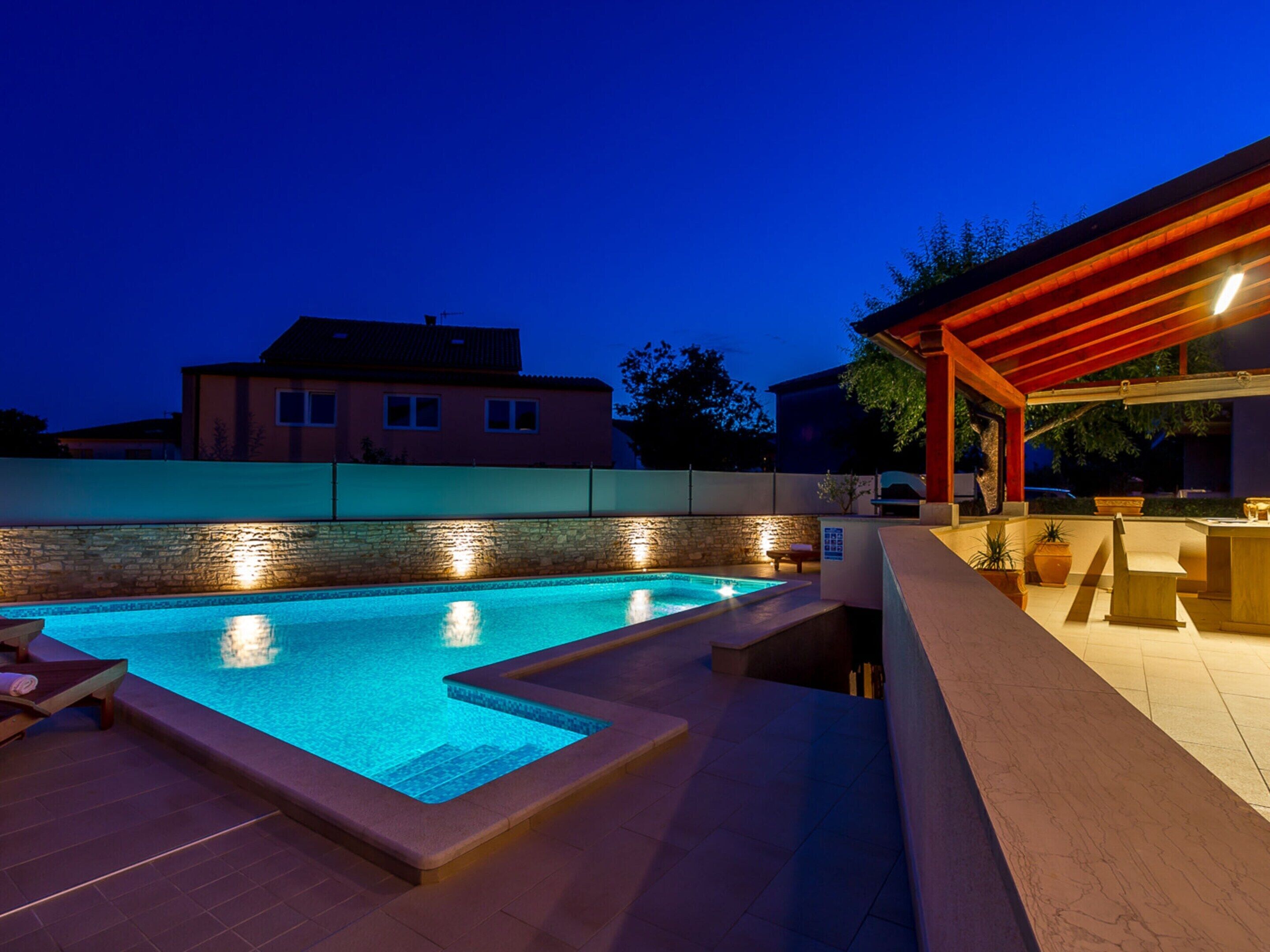 A swimming pool with a stone wall and a wooden deck, surrounded by a low wall and a house in the background.