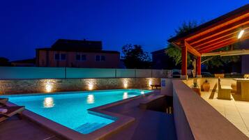 A swimming pool with a stone wall and a wooden deck, surrounded by a low wall and a house in the background.