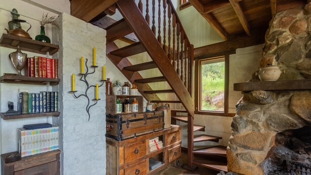 A rustic room with a stone fireplace, wooden staircase, and shelves with books and candles. - The Getaway Perth Hills (Roleystone)