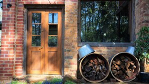 A brick building with a wooden double door and a window. There are two large metal barrels filled with logs and a potted plant near the window. - The Getaway Perth Hills (Roleystone)