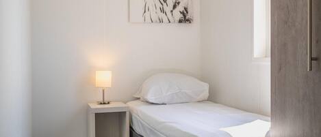 A small, neatly arranged bedroom with a single bed, a bedside table with a lamp, and a wall-mounted artwork.