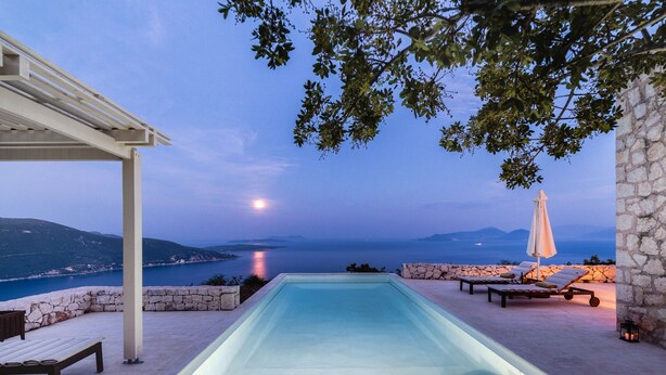 A swimming pool with a stone wall and lounge chairs, overlooking a body of water and hills. - Luxurious Villa in Vasiliki with Swimming Pool & Hot Tub (Lefkada)