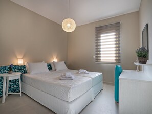 A bedroom with a large bed, bedside tables, a hanging light fixture, a window with blinds, and a potted plant on a side table.