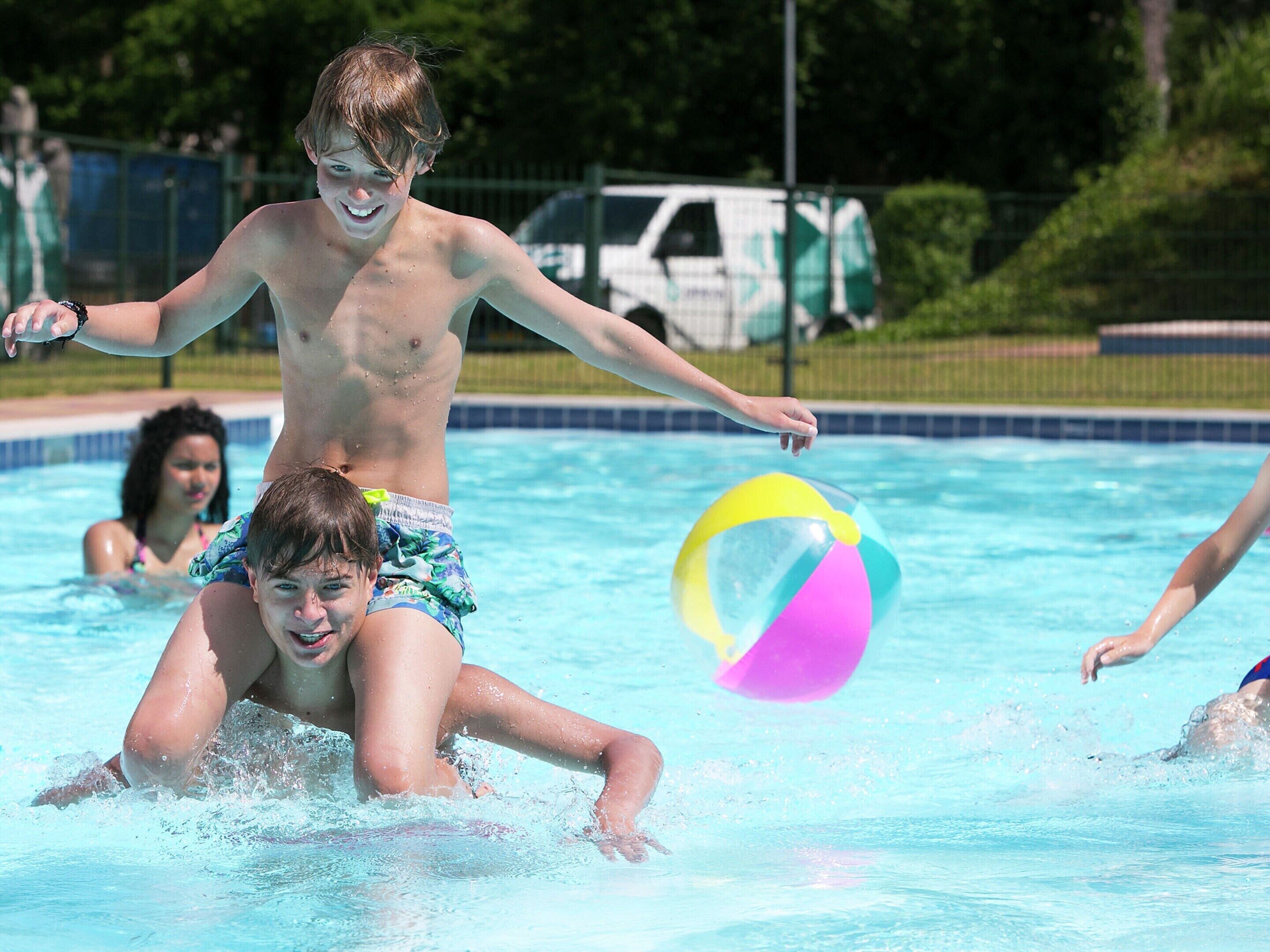 A swimming pool with clear blue water, a child playing with a ball, and a white van in the background.