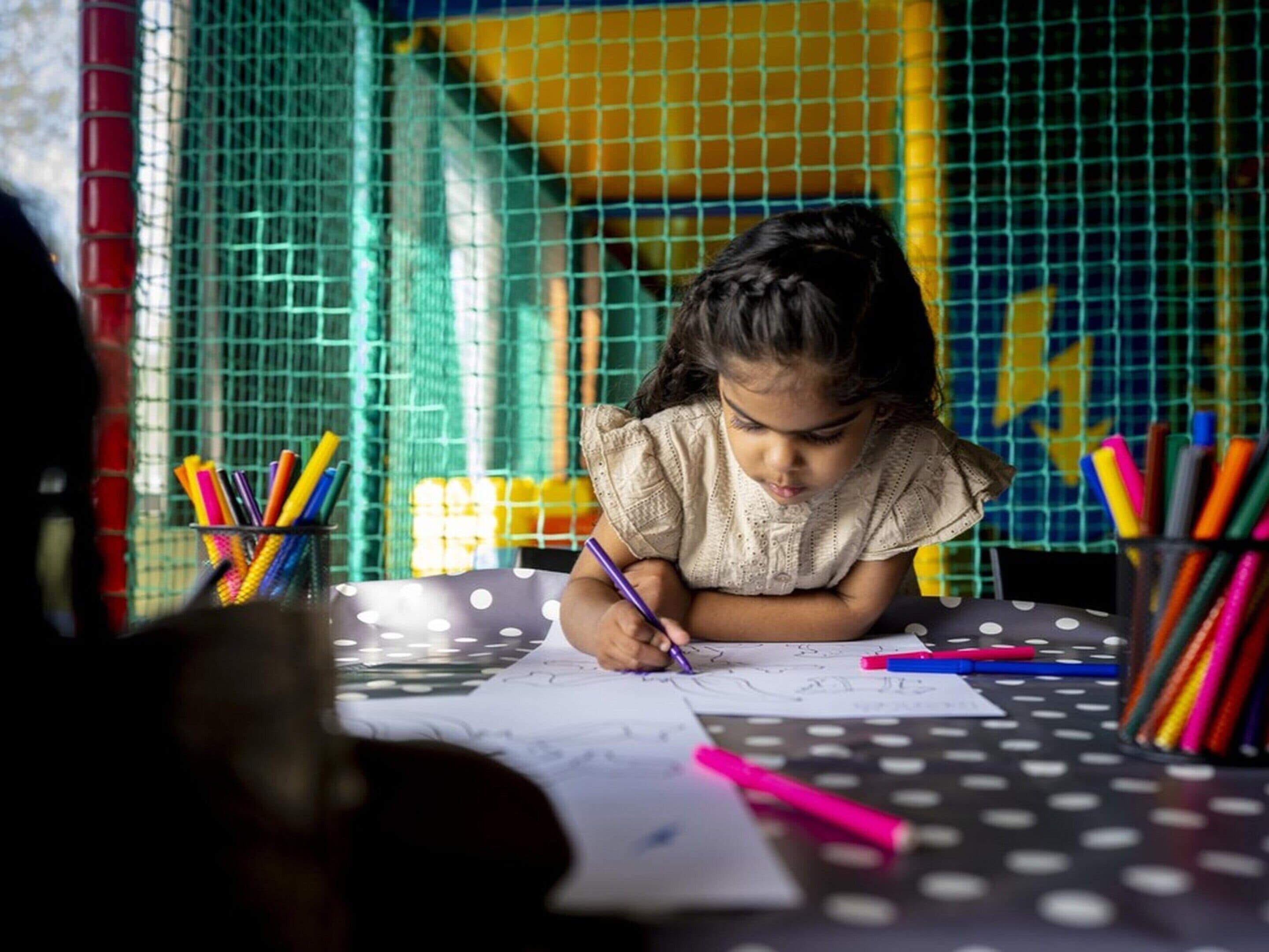 A child writing at a table with colorful pens and pencils, surrounded by a wire mesh and a yellow wall.