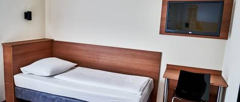 A hotel room with a single bed, a small desk, a chair, a flat-screen TV mounted on the wall, and a wall-mounted light fixture.